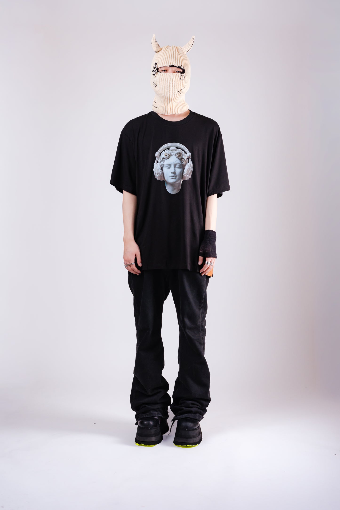 Person wearing a black t-shirt with a graphic design, black pants, and a white mask on a light gray background