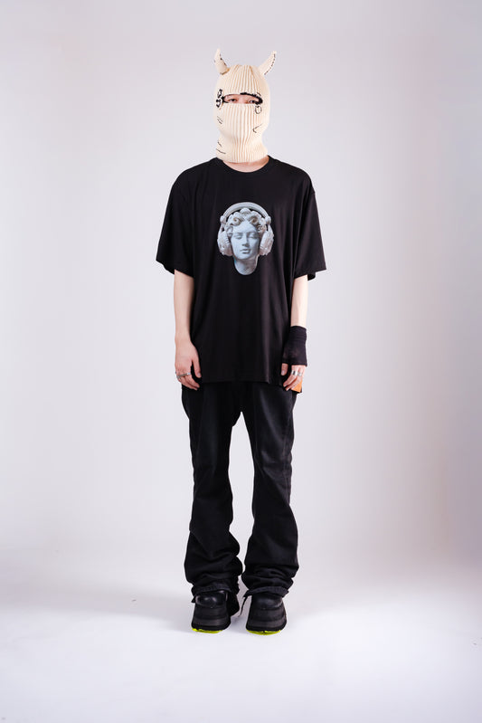 Person wearing a black t-shirt with a graphic design, black pants, and a white mask on a light gray background