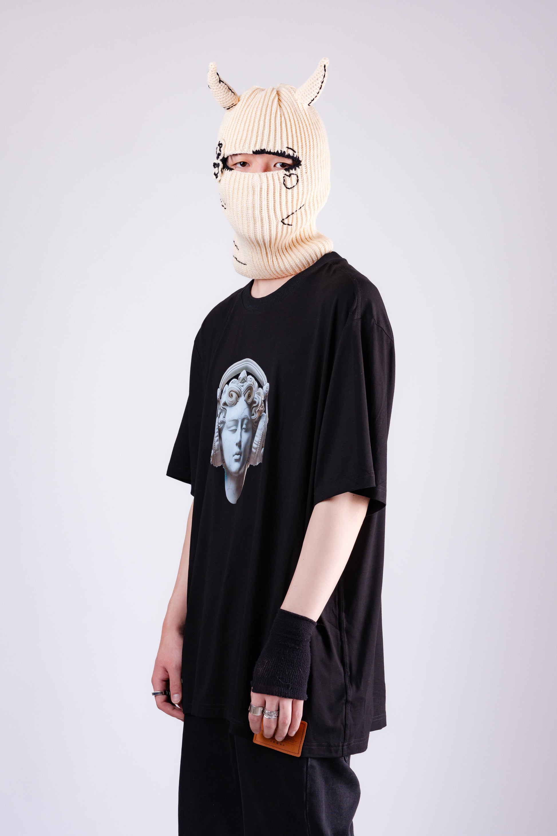 Person wearing a black oversized t-shirt with a graphic design, black pants, and a beige knit balaclava with animal ears on a white background.