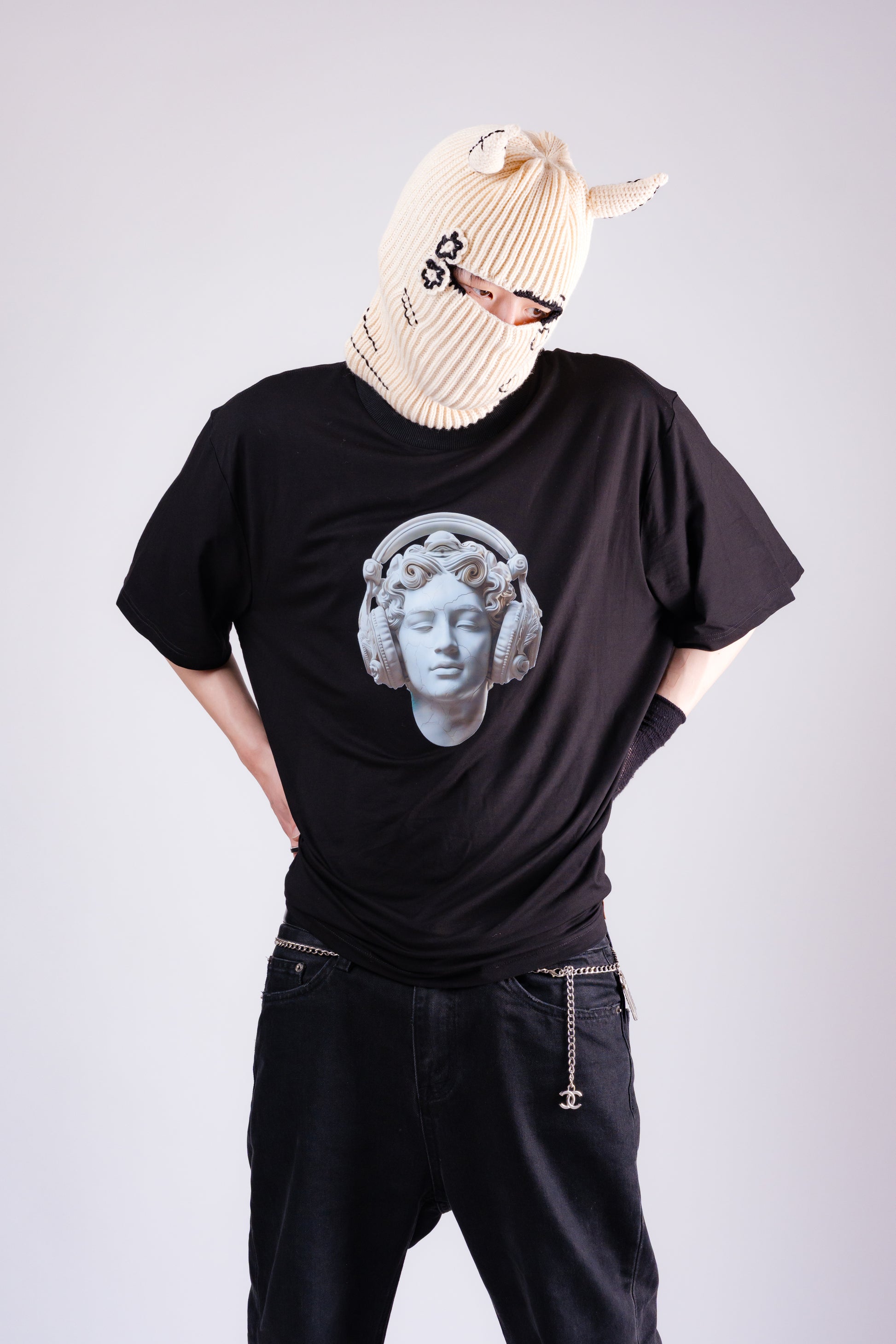 Person wearing a black t-shirt with a white graphic design, black pants, and a beige balaclava on a light gray background.