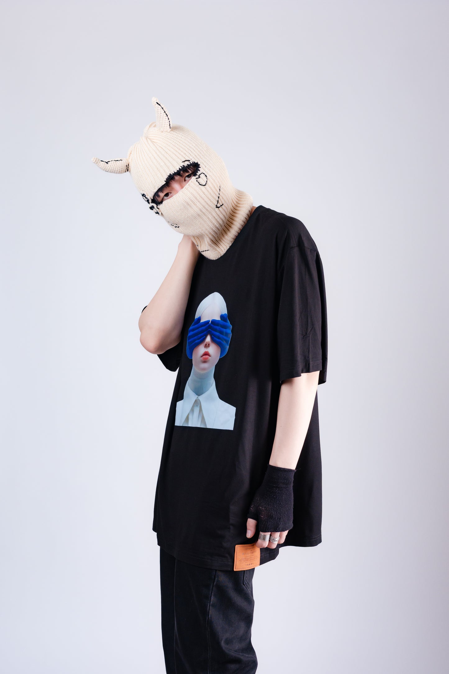 Person wearing a black t-shirt with a graphic design, black pants, and a beige knit hat on a light gray background.