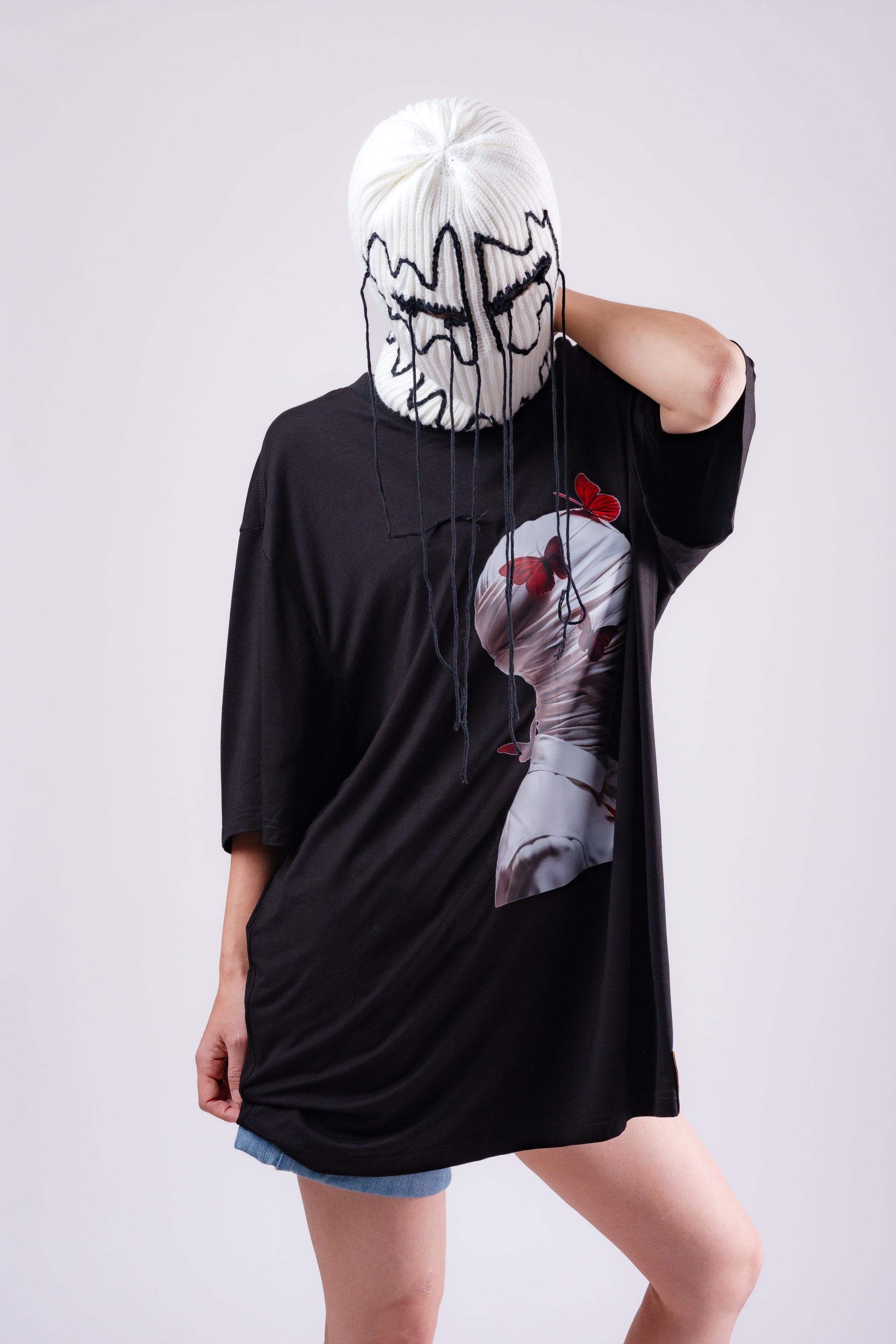 Person wearing a black oversized t-shirt with a graphic design and a white mask on a plain background